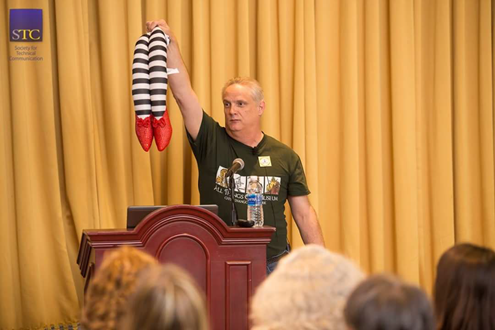 Presenter Ben Woelk holding a pair Wizard of Oz witch legs with stripes and ruby slippers, from STC Summit 2017 presentation, Follow the Yellow Brick Road: A Leadership Journey to the Emerald City